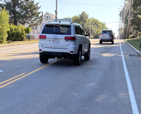 Cyclist's view of a two-way road with a two-way turn lane and narrow should with a crrb, cyclist riding in the right tire track, motorist straddle passing.